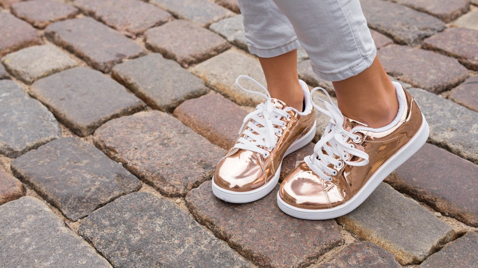 Close-up on a person's ankles and feet. They're wearing light-wash jeans and metallic gold sneakers and standing on a brick road.