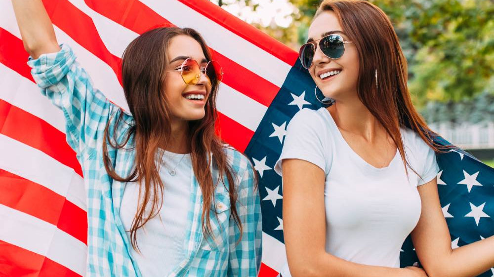 Two girls wearing festive Fourth of July clothing colors and holding the United States flag behind them