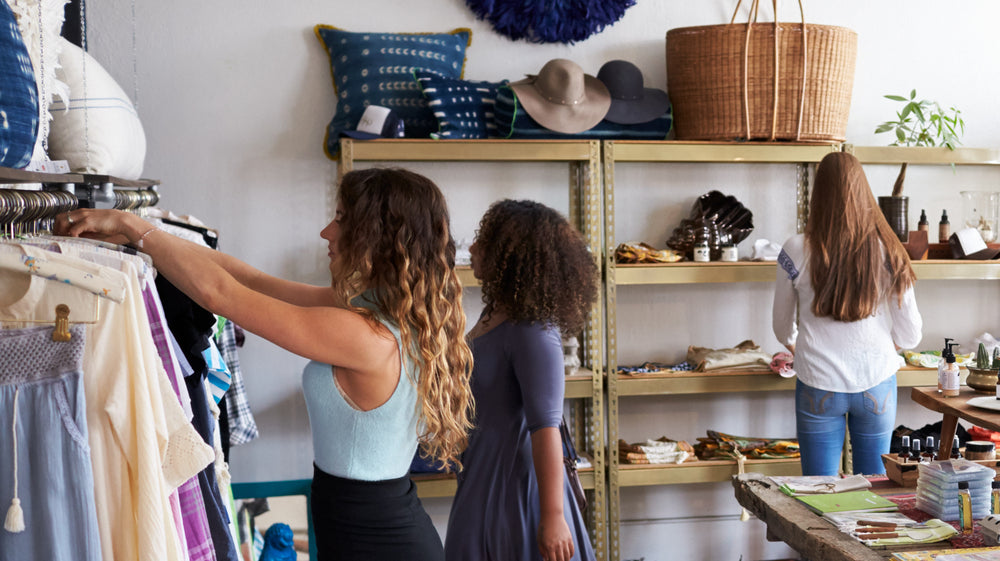Several customers shop in a busy clothing boutique, browsing tables and shelves lined with clothes and accessories.
