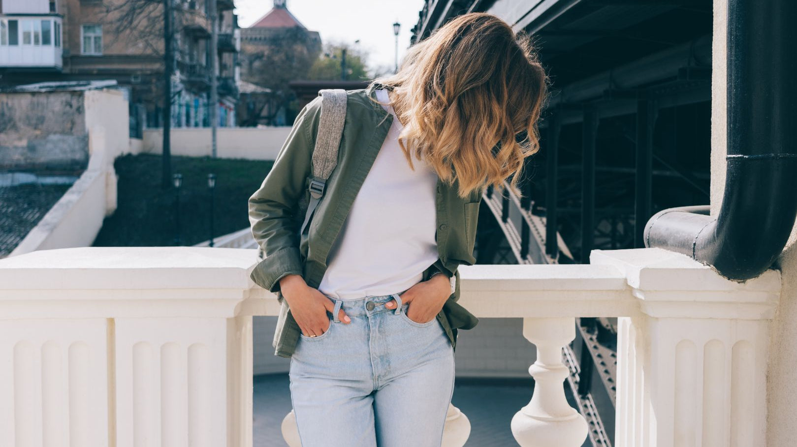 A young and stylish woman stands in front of a city landscape. She looks down, blonde hair obscuring her face.