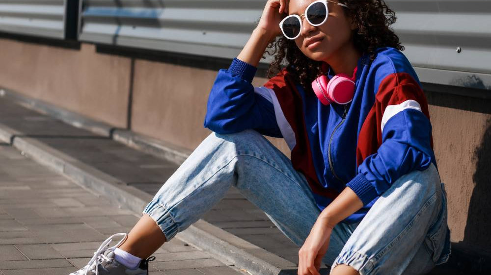 A girl wearing retro sunglasses, a bomber hoodie, high-waisted jeans, and other retro fashion styles sits on a curb.