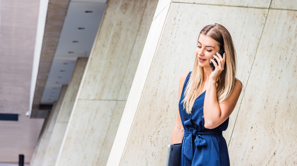 A young woman wearing a blue jumpsuit to the office, talking on the phone, and carrying a portable laptop.