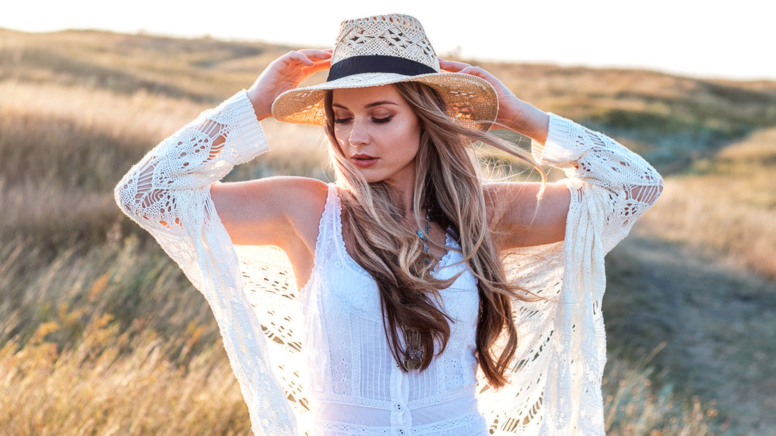 A woman wearing a summer cardigan and white dress stands in a field at sunset, holding onto her sunhat as the wind blows.