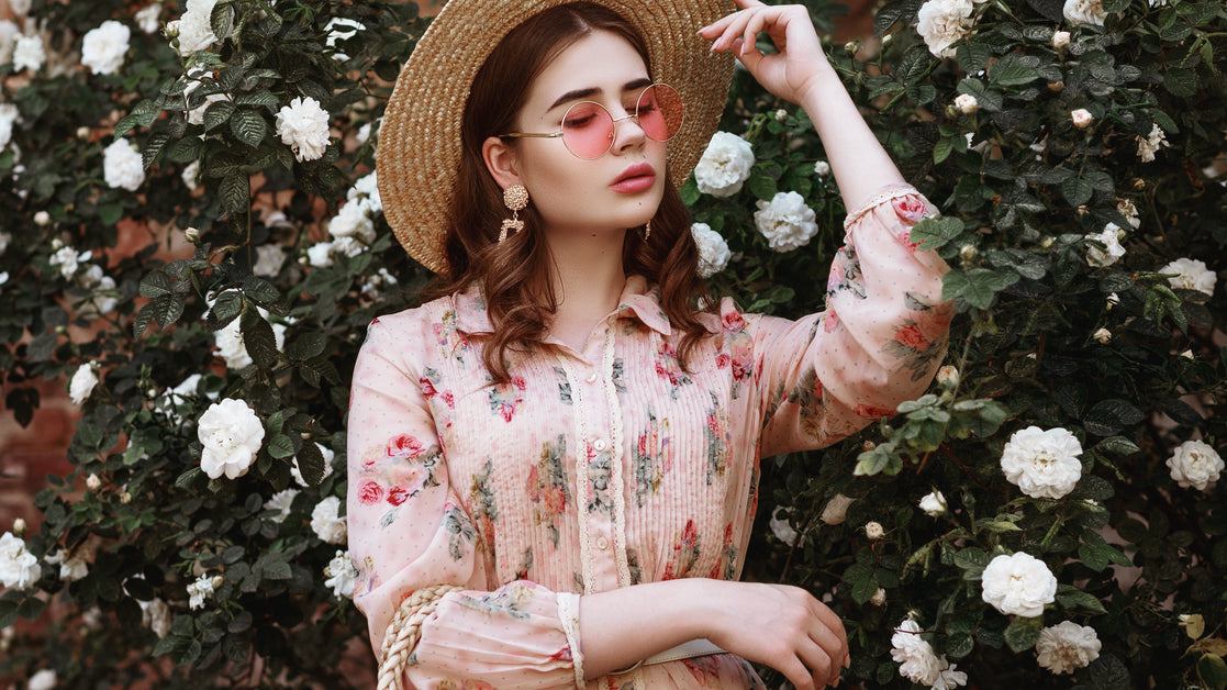 A brunette woman wearing a pink floral dress, pink sunglasses, and a straw hat stands in front of a white rose bush.