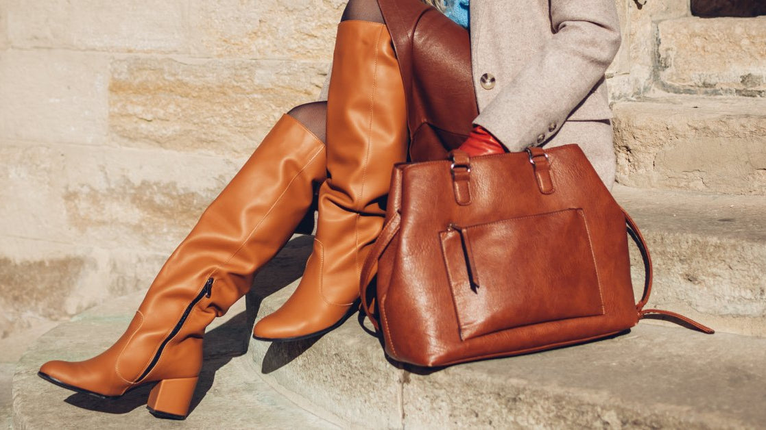 A close-up of a woman wearing orange leather boots, a leather skirt, and a beige coat while holding a leather bag.