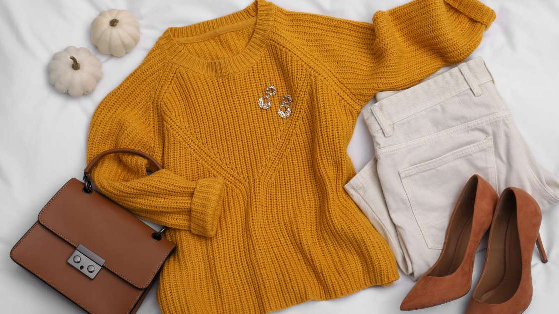 A fall outfit, including a mustard yellow sweater, off-white pants, stylish heels, and accessories, next to two small pumpkins.