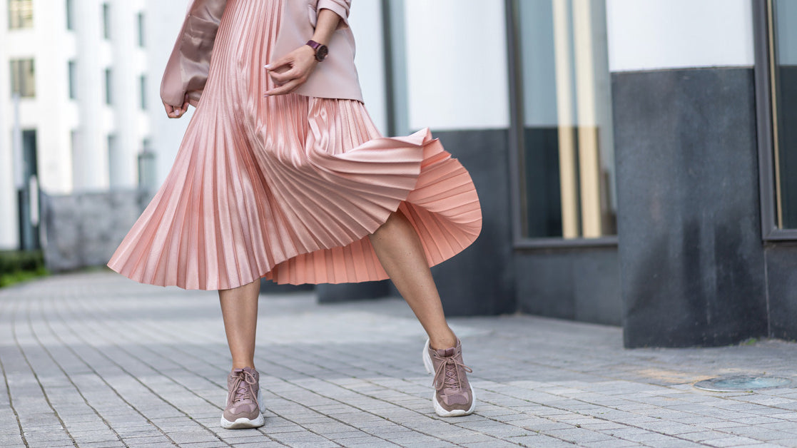 A close-up on a woman's outfit from the waist down. She wears a pastel pink pleated skirt, pink sneakers, and a pink coat.