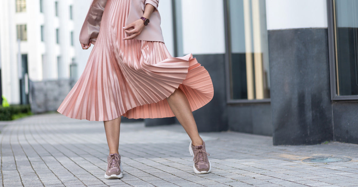 A close-up on a woman's outfit from the waist down. She wears a pastel pink pleated skirt, pink sneakers, and a pink coat.