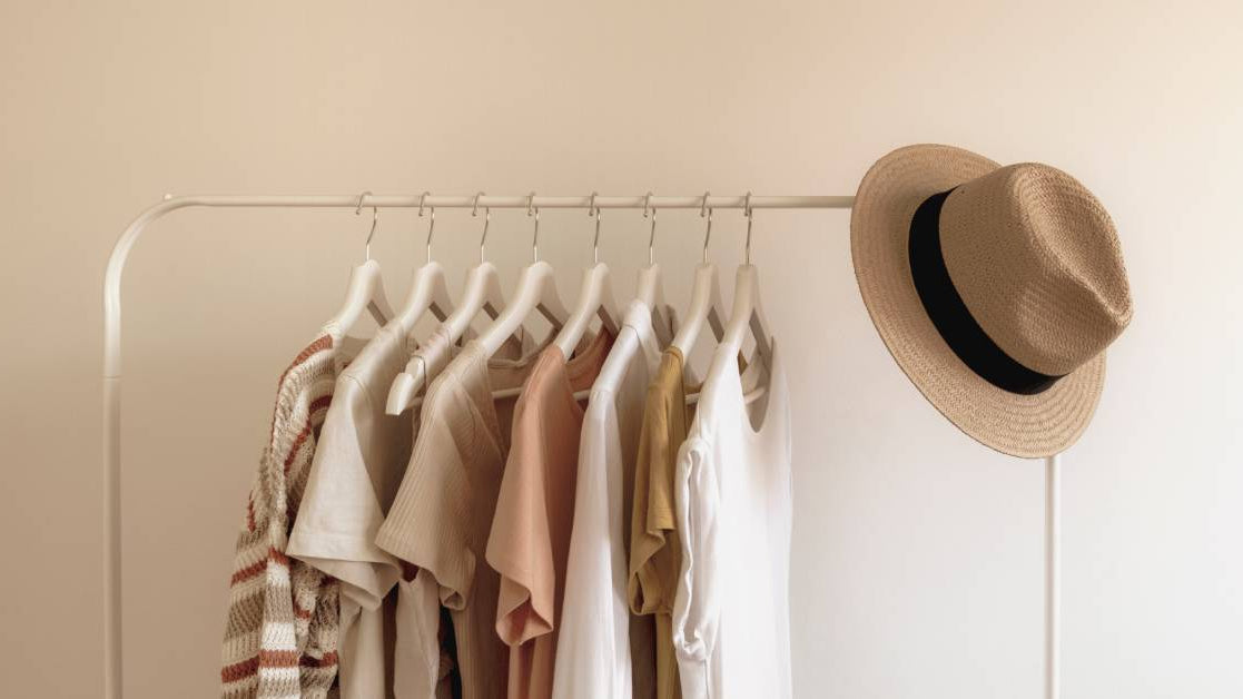 A row of neutral-colored T-shirts hangs on a clothing rack with a tan hat beside them, all set against a beige background.