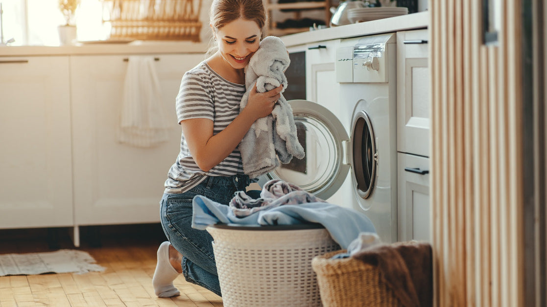 A woman kneels in her laundry room in front of a dryer, unloading clothes and holding a fluffy garment up to her face.