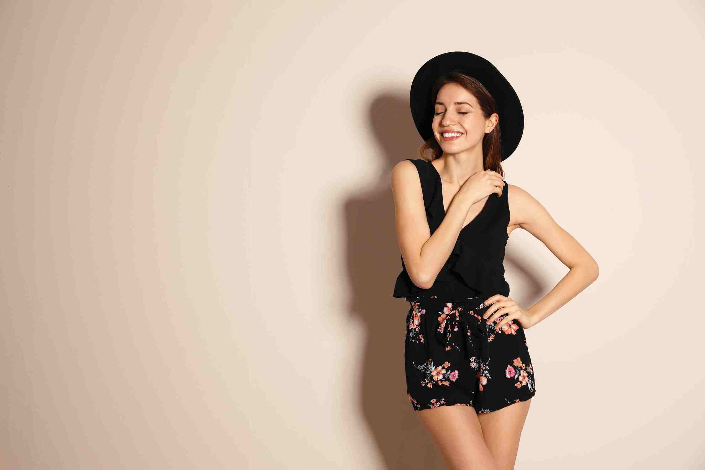 A young woman smiling and wearing black shorts with a floral pattern, a black tank top, and a black hat