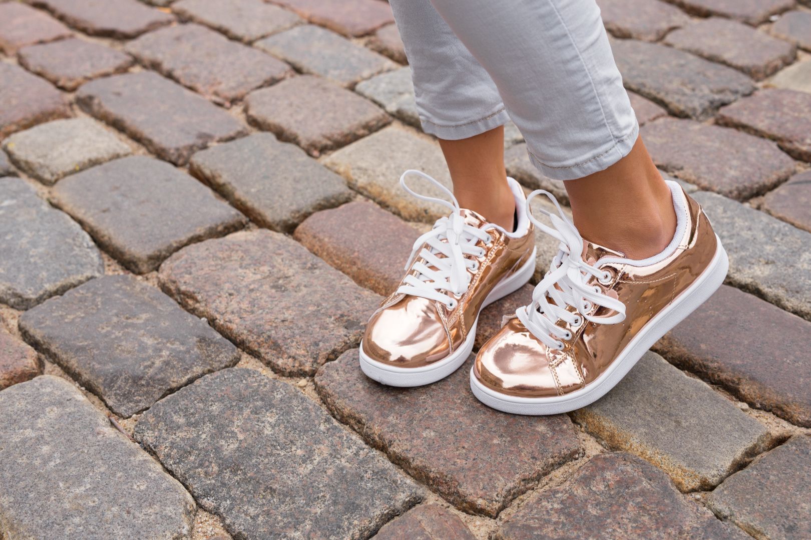 Close-up on a person's ankles and feet. They're wearing light-wash jeans and metallic gold sneakers and standing on a brick road.