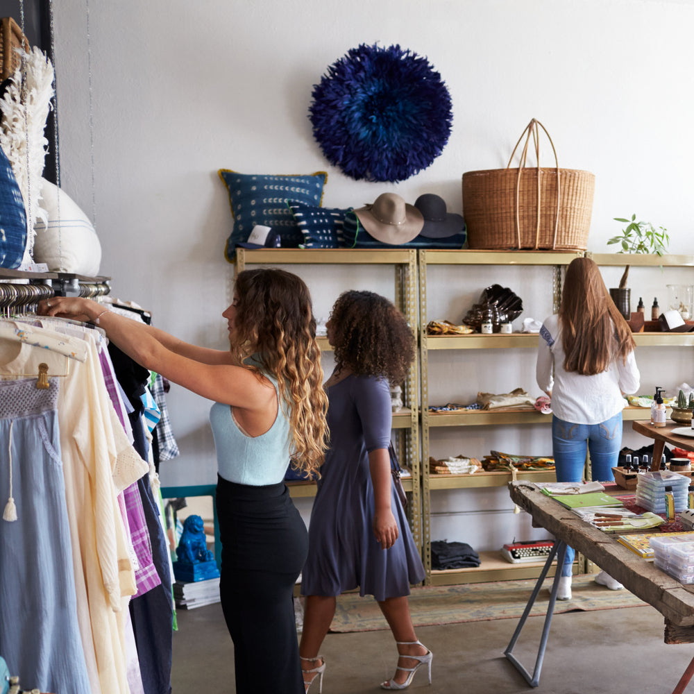 Several customers shop in a busy clothing boutique, browsing tables and shelves lined with clothes and accessories.