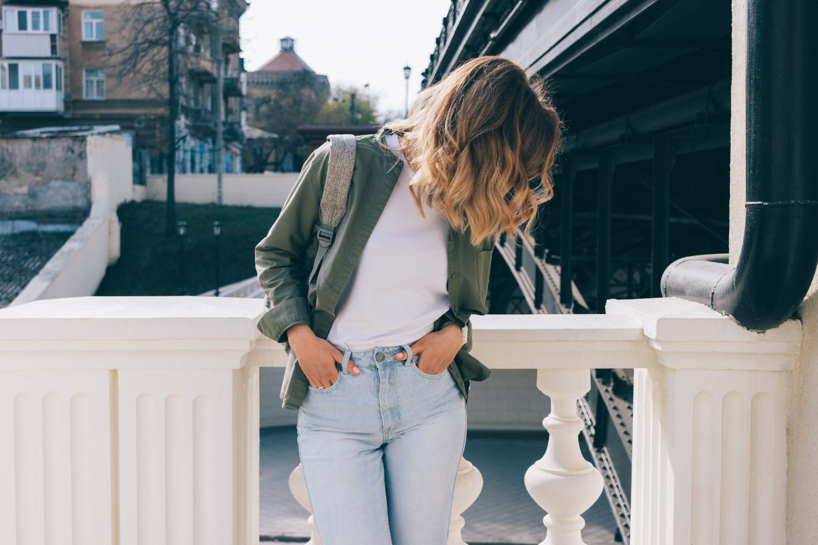 A young and stylish woman stands in front of a city landscape. She looks down, blonde hair obscuring her face.