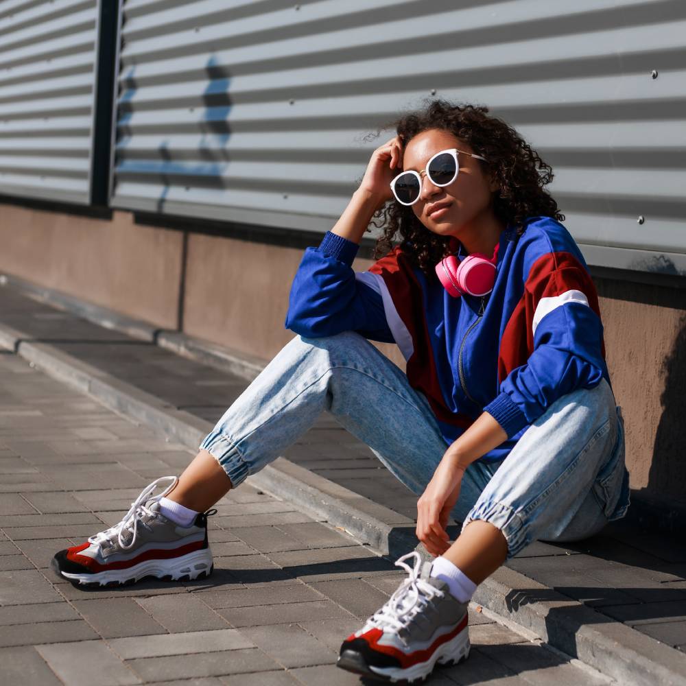  A girl wearing retro sunglasses, a bomber hoodie, high-waisted jeans, and other retro fashion styles sits on a curb.