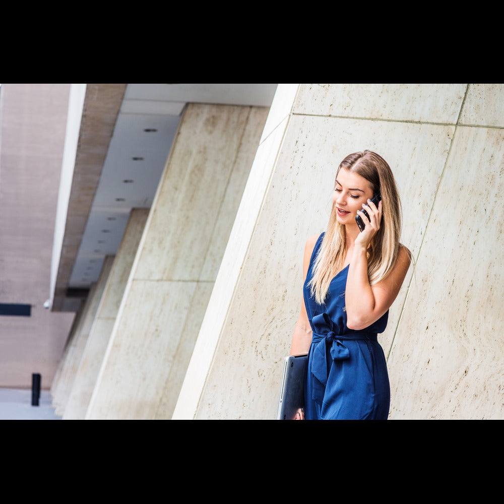 A young woman wearing a blue jumpsuit to the office, talking on the phone, and carrying a portable laptop.