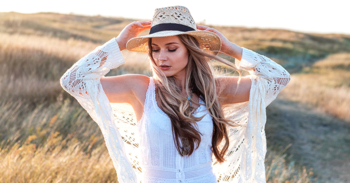 A woman wearing a summer cardigan and white dress stands in a field at sunset, holding onto her sunhat as the wind blows.