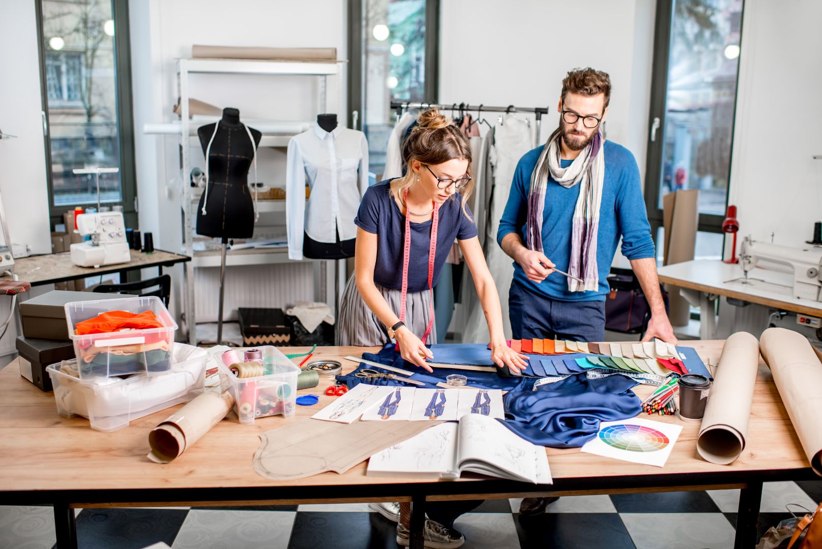 Two fashion designers stand at a worktable, spreading out various fabric swatches and materials for clothing design.