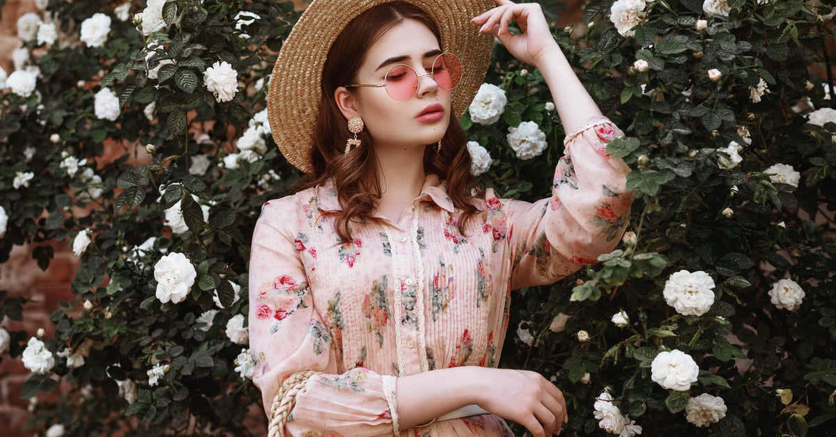 A brunette woman wearing a pink floral dress, pink sunglasses, and a straw hat stands in front of a white rose bush.