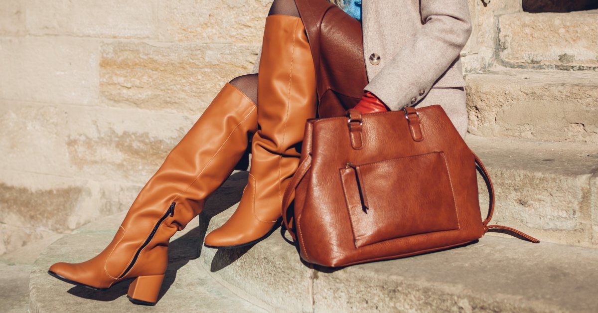 A close-up of a woman wearing orange leather boots, a leather skirt, and a beige coat while holding a leather bag.