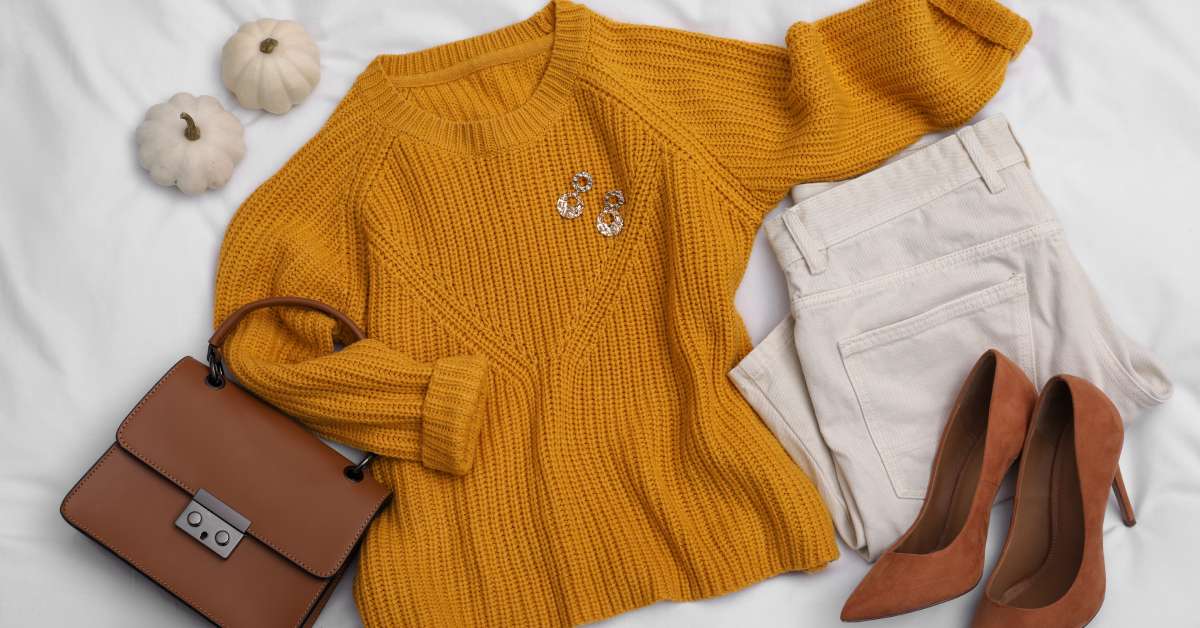 A fall outfit, including a mustard yellow sweater, off-white pants, stylish heels, and accessories, next to two small pumpkins.