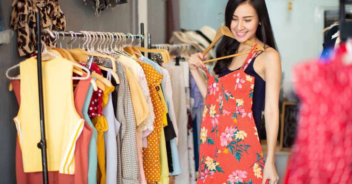 A young woman holds up a red floral dress to her figure as she browses a clothing rack in a boutique.