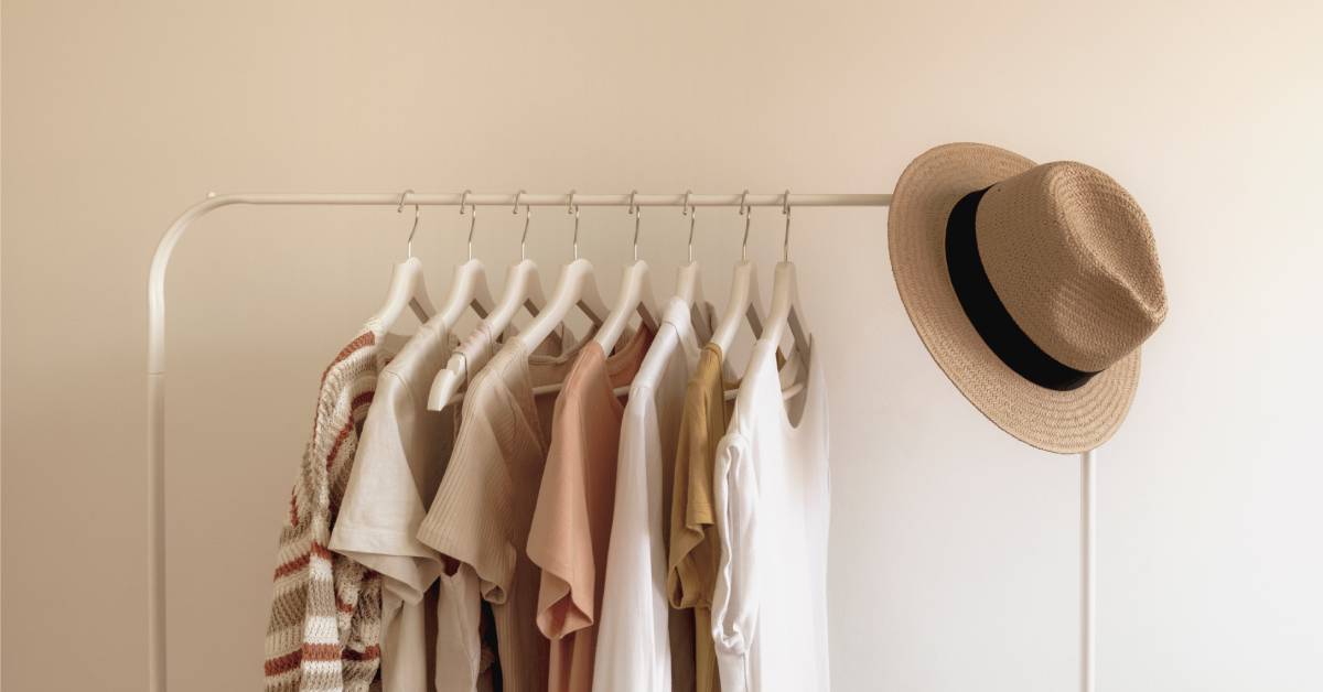 A row of neutral-colored T-shirts hangs on a clothing rack with a tan hat beside them, all set against a beige background.