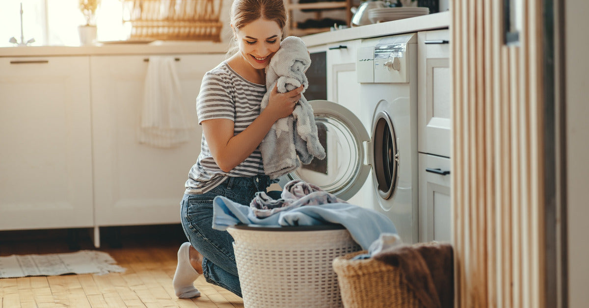 A woman kneels in her laundry room in front of a dryer, unloading clothes and holding a fluffy garment up to her face.